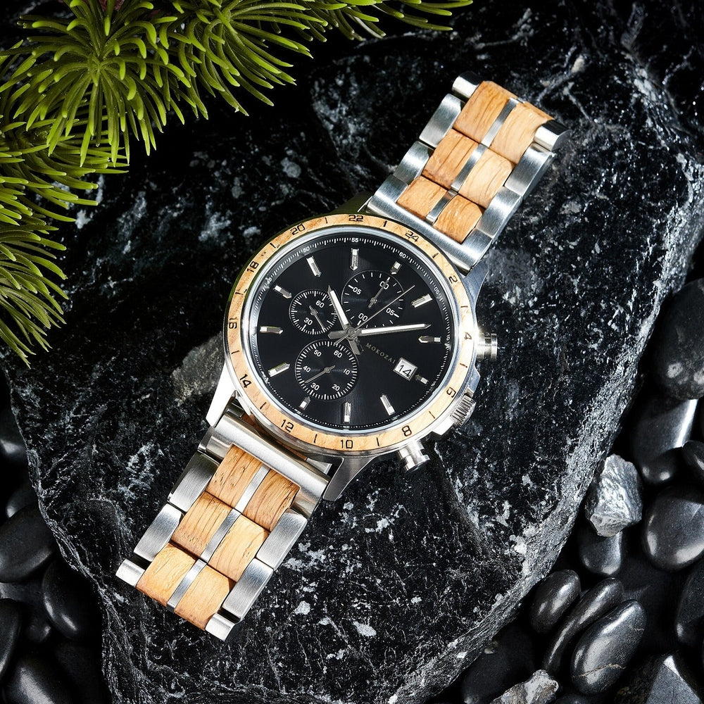 Mokozai men's wooden and stainless steel watch with a natural wood inlay, black dial, and chronograph functions, resting on a sleek black stone surface with green pine branches. This stylish eco-friendly timepiece combines nature-inspired materials with modern craftsmanship, perfect for environmentally conscious customers in Australia.