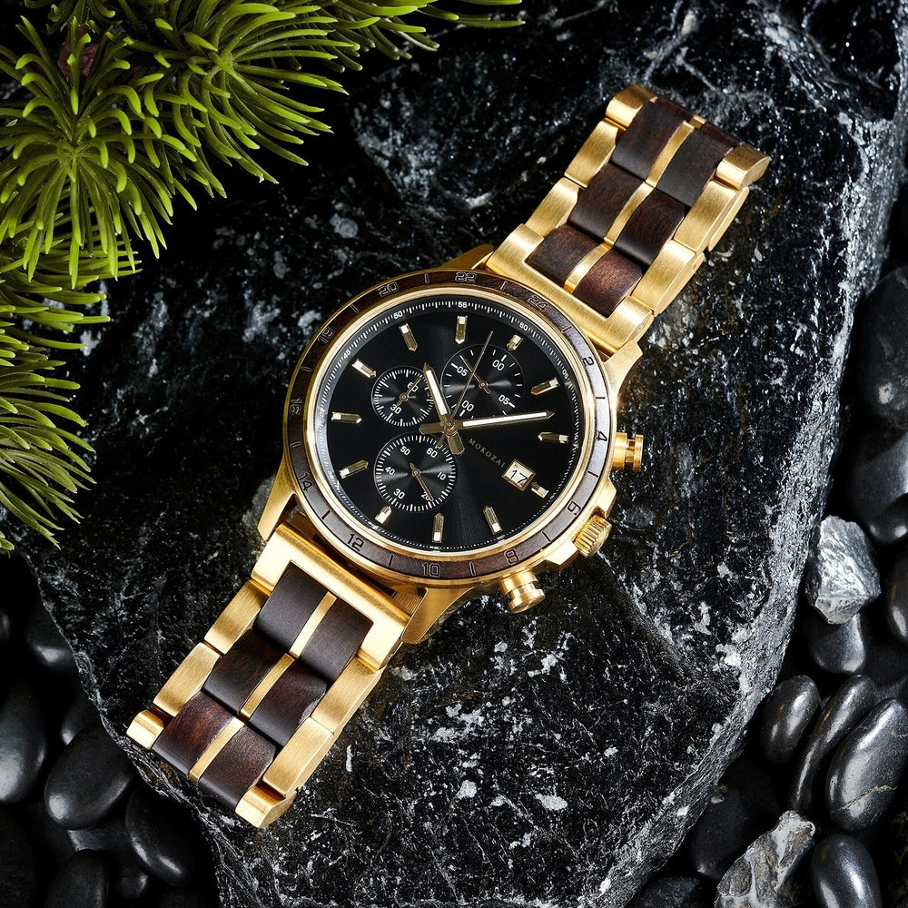 Elegant gold wristwatch with a black face and gold accents, featuring a black and gold link bracelet, displayed on a dark stone background with greenery in the corner.