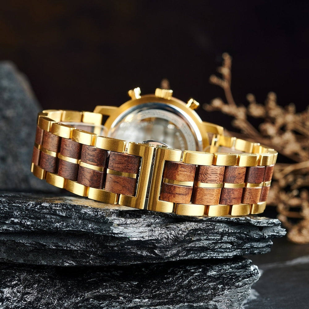 Close-up of a luxury wooden and gold wristwatch, featuring an intricate link bracelet with wood inlays and a polished metal clasp, beautifully set on layered black stones with dried decorative foliage in the background.