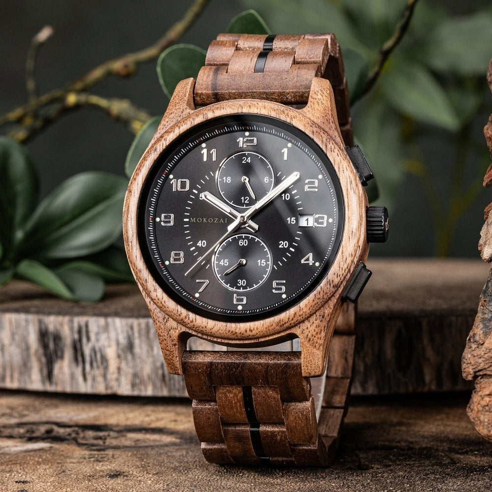 A wooden wristwatch featuring a black dial with white numerals and sub-dials, paired with a natural wooden case and matching wood link bracelet, set against a rustic backdrop with green leaves.