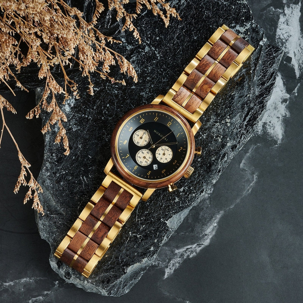 Luxury wooden and gold metal wristwatch with a black dial and chronograph, elegantly displayed on a dark stone surface, alongside delicate dried foliage for a sophisticated and natural aesthetic.