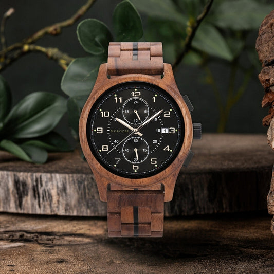 Stylish wooden wristwatch with a dark dial, chronograph features, and luminous hands, complemented by a wooden link bracelet. The watch is elegantly displayed on a rustic wooden surface with natural green leaves in the background.